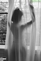 A woman standing in front of a window with sheer curtains.