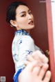 A woman in a blue and white cheongsam leaning against a red door.