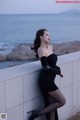 A woman in a black dress leaning against a wall by the ocean.